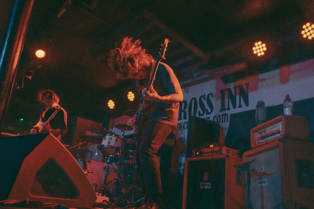 A guitarist with long hair plays energetically on stage under red lights, while a drummer and another musician perform beside him. Amplifiers, drums, and a banner reading ROSS INN are visible in the background.