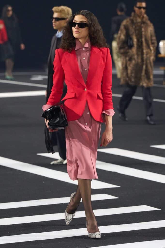 A model walks on a runway wearing a bright red blazer over a pale pink dress, with sheer black tights, snakeskin-patterned heels, sunglasses, and carrying a black clutch. Other models are visible in the background.