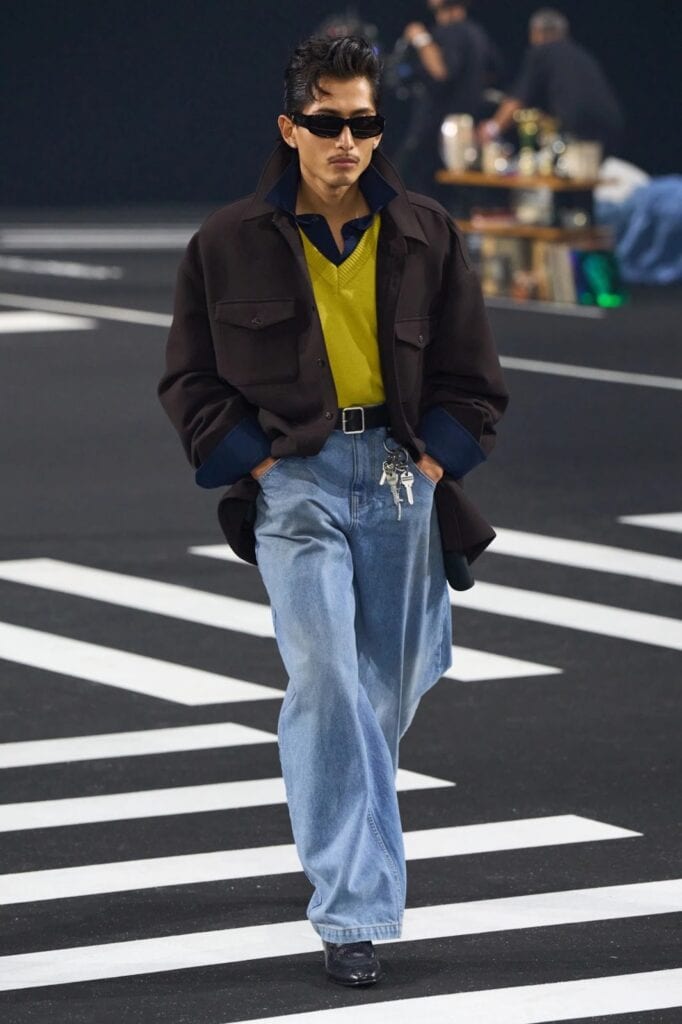 A model walks on a runway wearing a dark jacket over a yellow shirt with a blue collar, wide-legged light blue jeans with a keychain accessory, black shoes, and dark sunglasses. The background is a street-style set.