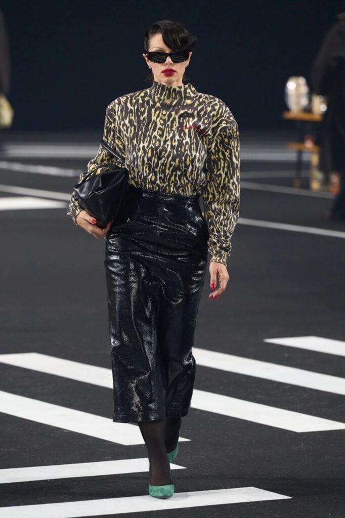 A model walks on a runway wearing a leopard print turtleneck blouse, a shiny black midi skirt, black tights, green shoes, dark sunglasses, and carries a large black clutch.