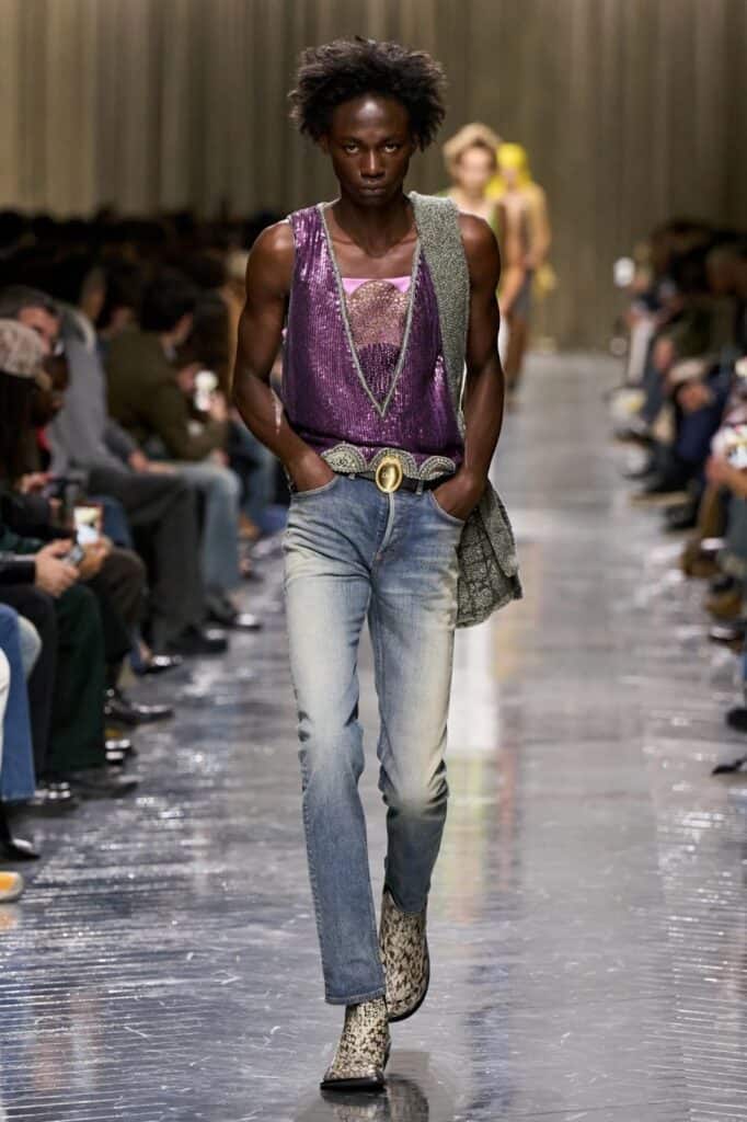 A model walks down a runway wearing a shiny purple sleeveless top, light blue jeans, a large textured belt, a gray bag slung over one shoulder, and patterned boots. The audience sits on both sides of the glossy runway.