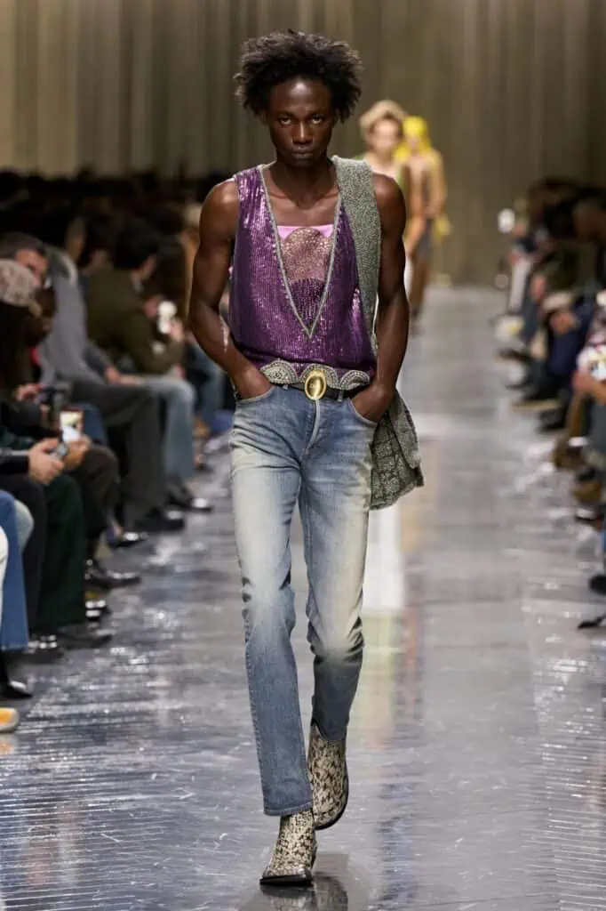 A model walks down a runway wearing a shiny purple sleeveless top, light blue jeans, a large textured belt, a gray bag slung over one shoulder, and patterned boots. The audience sits on both sides of the glossy runway.