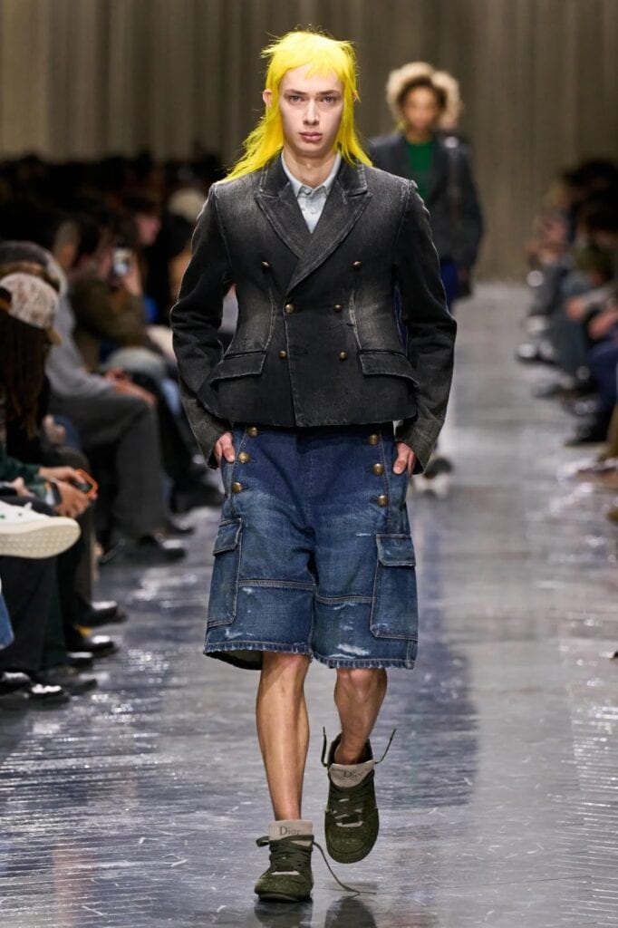 A model with bright yellow hair walks a runway wearing a dark double-breasted blazer, denim cargo shorts, and green slip-on shoes. The audience is seated on both sides of the reflective runway.