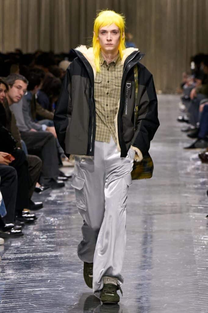A model with bright yellow hair walks down a fashion runway wearing a black and gray jacket, a beige plaid shirt, shiny silver cargo pants, and dark green shoes. The audience sits on either side of the runway.