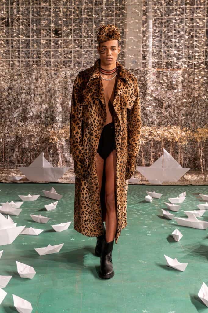 A person wearing a long leopard print coat, black boots, and a matching hat walks on a green floor covered with white paper boats, set against a textured silver and gold backdrop.