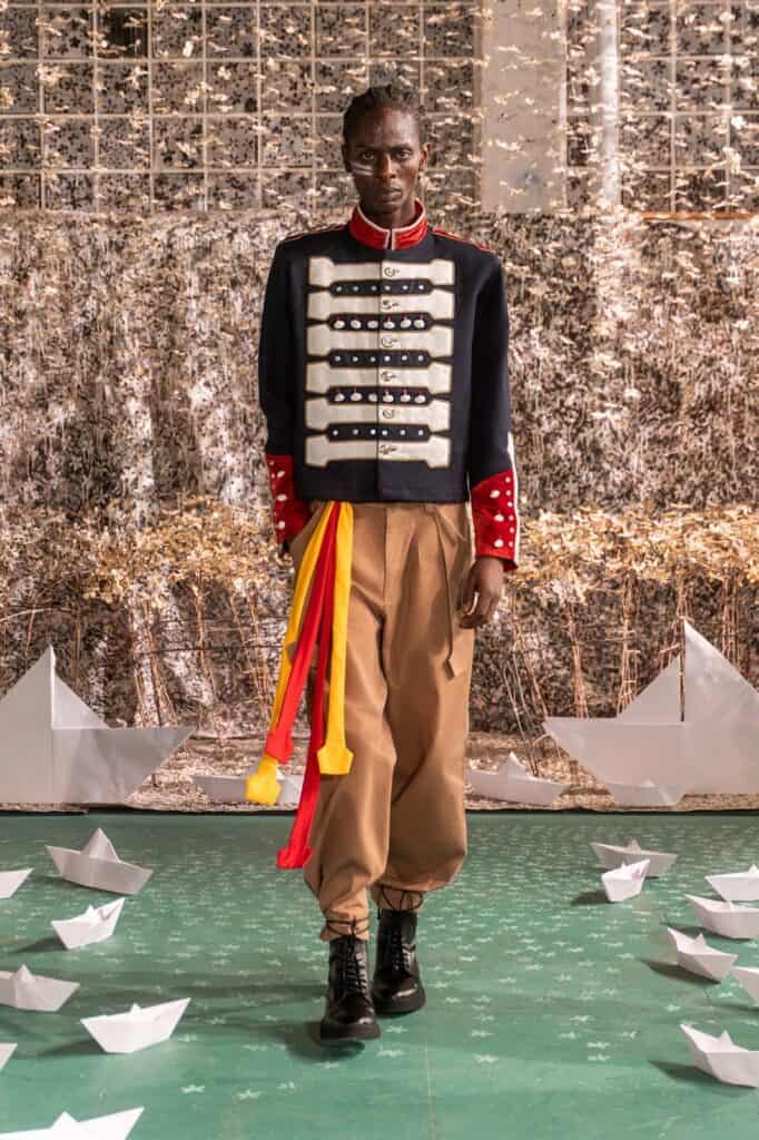 A model walks a runway wearing a navy military-style jacket with decorative buttons, tan high-waisted pants, black boots, and colorful ribbons; paper boats are scattered on the green floor, with a shimmering gold backdrop.