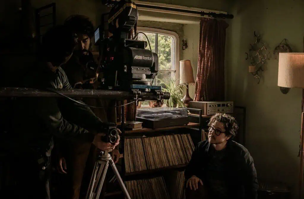 Two people operate a large camera on a tripod indoors while another person with glasses sits nearby. They are in a cozy, dimly lit room with records, bookshelves, and a window letting in natural light.