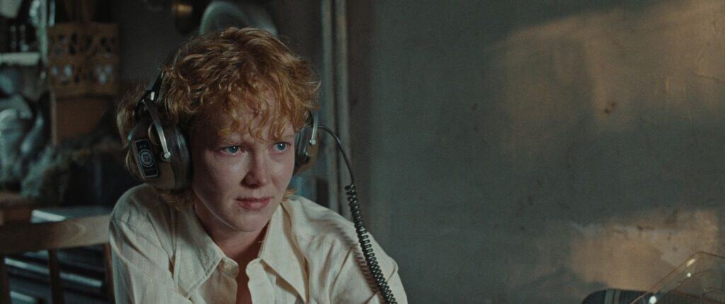 A person with short curly blond hair wearing large headphones and a light-colored shirt sits indoors, looking serious. There is a shadowy wall in the background and sunlight coming through a window.