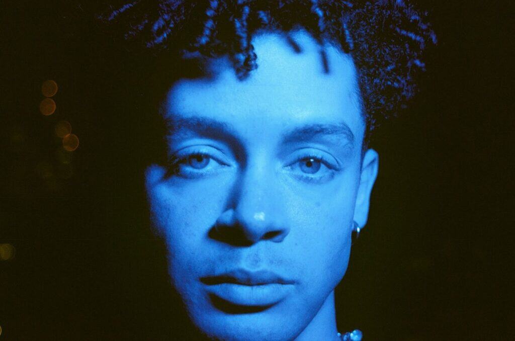 A close-up of a person with short curly hair and an earring, illuminated by blue lighting, looking directly at the camera with a neutral expression. The background is dark and out of focus.
