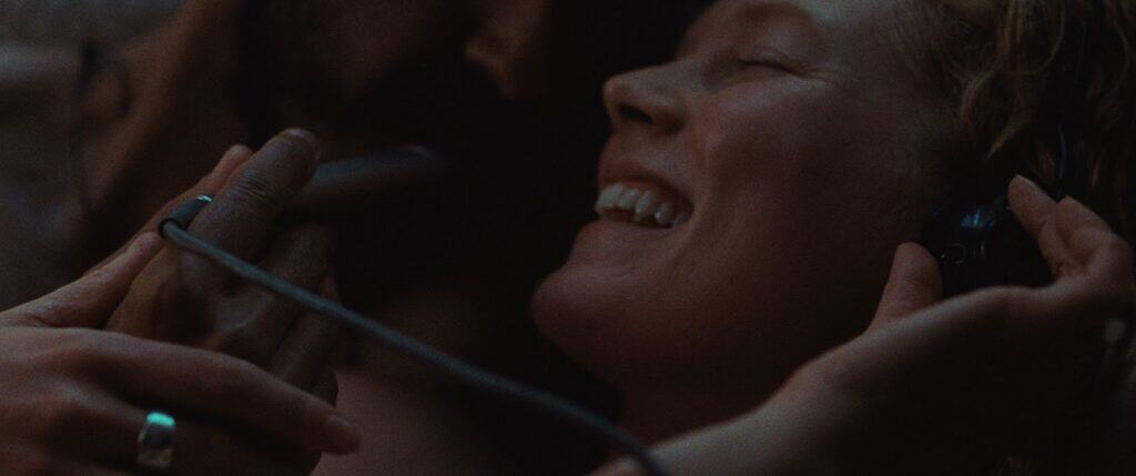 A close-up of a smiling woman with headphones on, eyes closed, enjoying music while another person gently holds the headphones and a microphone near her face in a dimly lit setting.