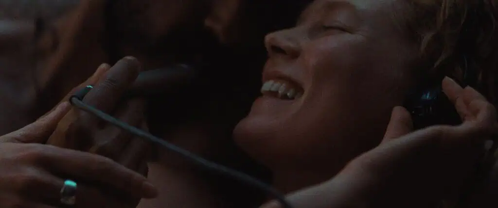 A close-up of a smiling woman with headphones on, eyes closed, enjoying music while another person gently holds the headphones and a microphone near her face in a dimly lit setting.