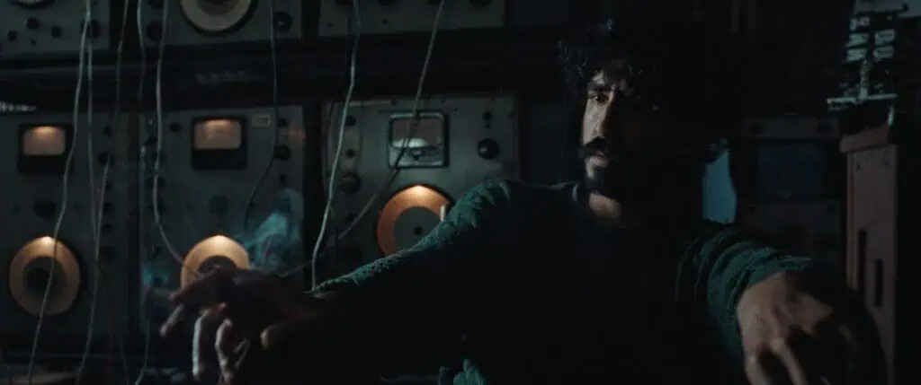 A man with dark curly hair and a beard sits in a dimly lit room, reaching out with his hands. Behind him are vintage audio equipment and machines with glowing dials and spools, suggesting a retro or industrial setting.