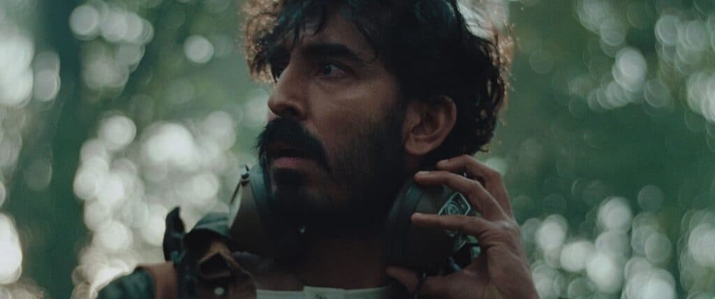 A man with a dark beard and mustache looks to the side with a concerned expression, holding large headphones around his neck. The background is blurred with green and light bokeh, suggesting an outdoor forest setting.