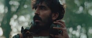 A man with a dark beard and mustache looks to the side with a concerned expression, holding large headphones around his neck. The background is blurred with green and light bokeh, suggesting an outdoor forest setting.