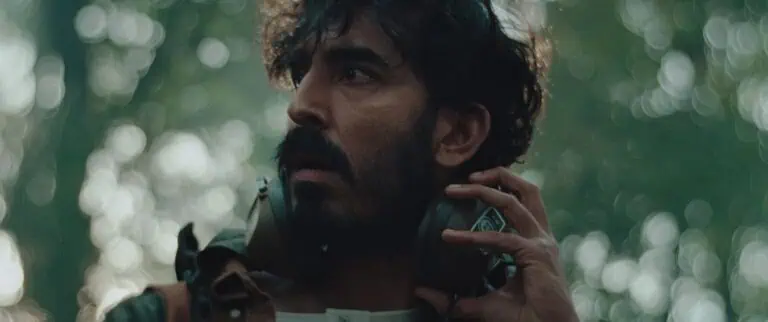 A man with a dark beard and mustache looks to the side with a concerned expression, holding large headphones around his neck. The background is blurred with green and light bokeh, suggesting an outdoor forest setting.