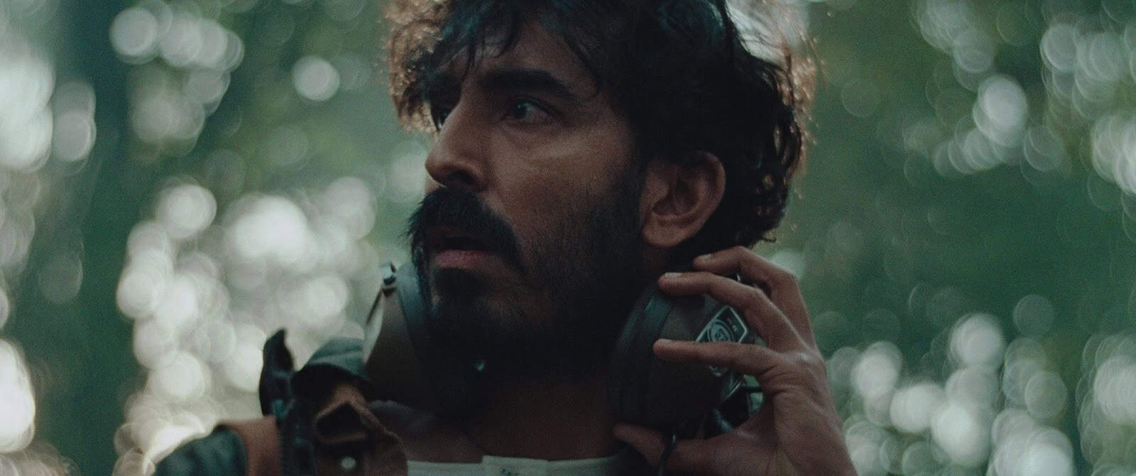 A man with a dark beard and mustache looks to the side with a concerned expression, holding large headphones around his neck. The background is blurred with green and light bokeh, suggesting an outdoor forest setting.