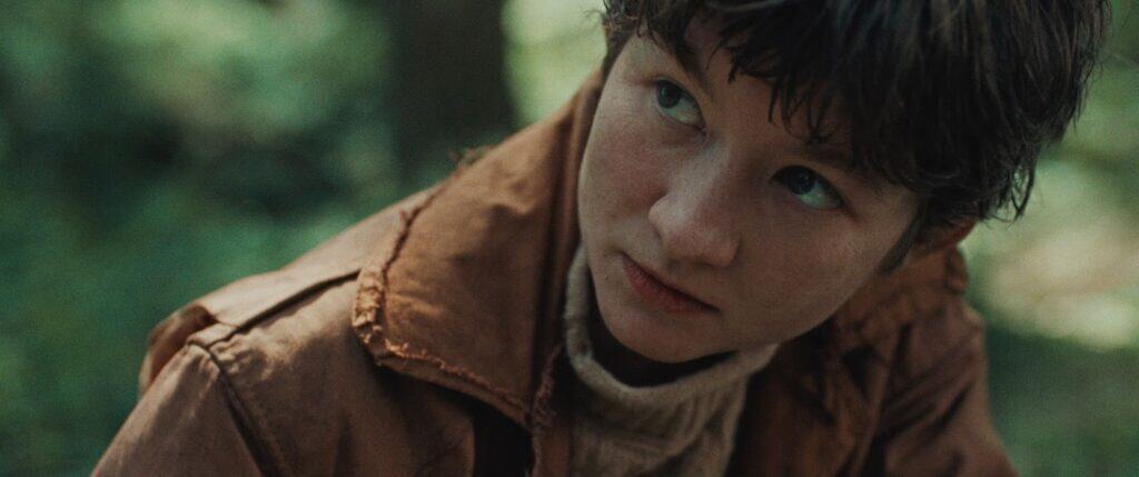 A young person with short brown hair wearing a brown jacket and beige turtleneck looks upward with a serious expression in an outdoor, forest setting.