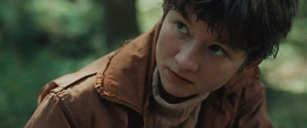 A young person with short brown hair wearing a brown jacket and beige turtleneck looks upward with a serious expression in an outdoor, forest setting.