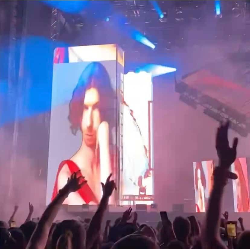 A crowd at a concert raises their hands while a large digital screen on stage displays a person in a red outfit, with blue and purple lighting effects illuminating the scene.