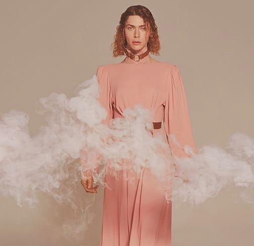 A person with shoulder-length curly hair wears a long, flowing peach dress and a choker necklace, standing against a neutral background with clouds of white smoke surrounding their waist.