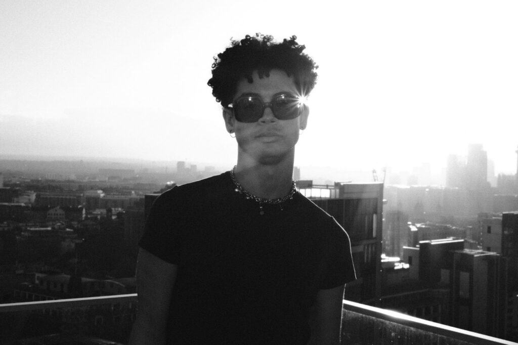 A person with curly hair and sunglasses stands on a rooftop at sunset, with city buildings and a hazy skyline in the background. Bright sunlight creates a lens flare over one lens of the sunglasses.