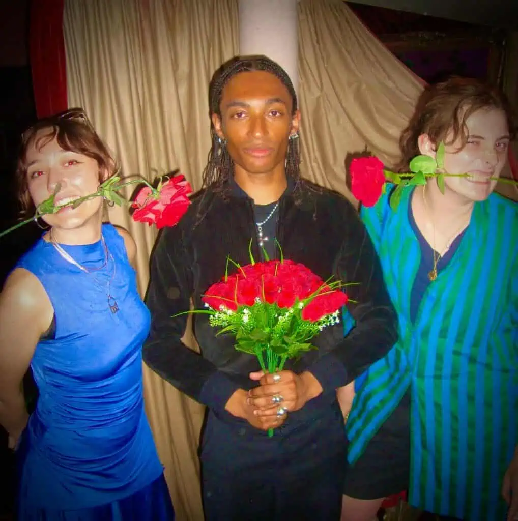 Three people pose indoors; the person in the center holds a bouquet of red roses, while the two on either side hold single roses in their mouths. Curtains hang in the background, and all three wear vibrant clothing and jewelry.