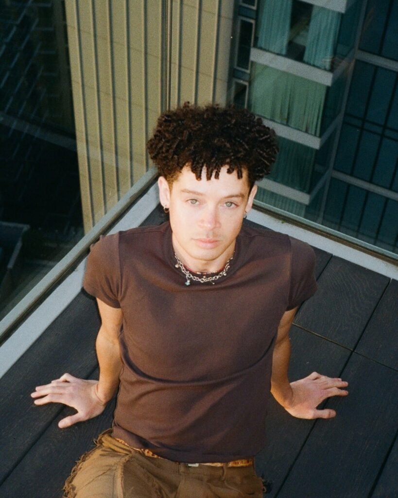 A young man with curly hair and a beaded necklace sits on a balcony deck, leaning back on his hands, with tall city buildings reflected in the glass behind him.