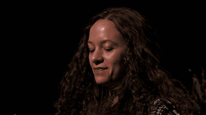 Jeda Pearl looks downward with a gentle expression, set against a dark background with part of a plant visible on the right.
