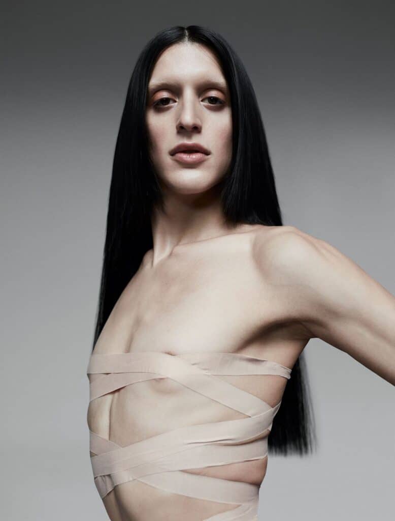 A person with long, straight black hair poses against a neutral background, wearing nude-colored fabric strips wrapped around their chest and torso. They have a calm expression and are looking directly at the camera.