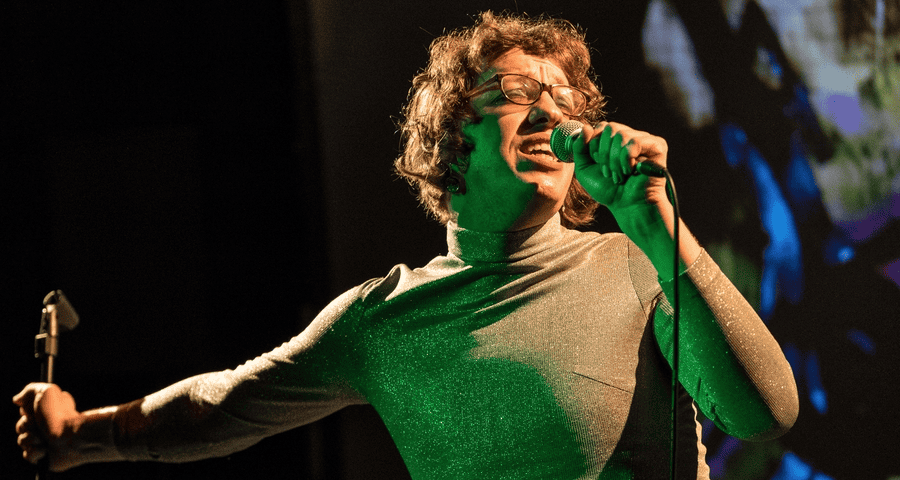 Harry Josephine Giles with short curly hair and glasses passionately sings into a microphone on stage, wearing a shimmery long-sleeve top, with dramatic lighting and a colorful background.