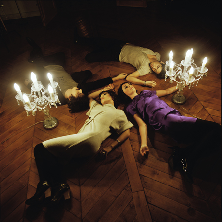 Four people lie on a wooden floor in a circular arrangement, heads close together, next to two ornate candelabras with lit candles. The scene is dimly lit and artistic.