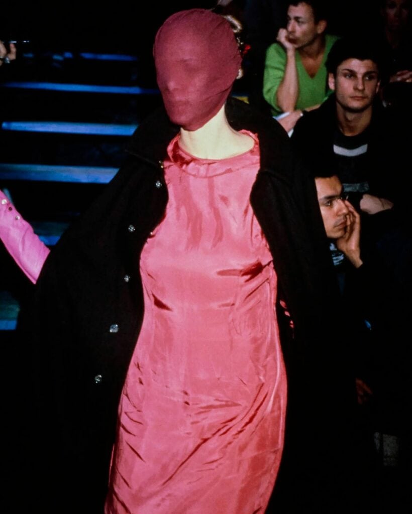 A model walks on a runway wearing a shiny pink dress and a matching pink fabric mask covering their entire head, with a black coat draped over their shoulders. Audience members watch in the background.