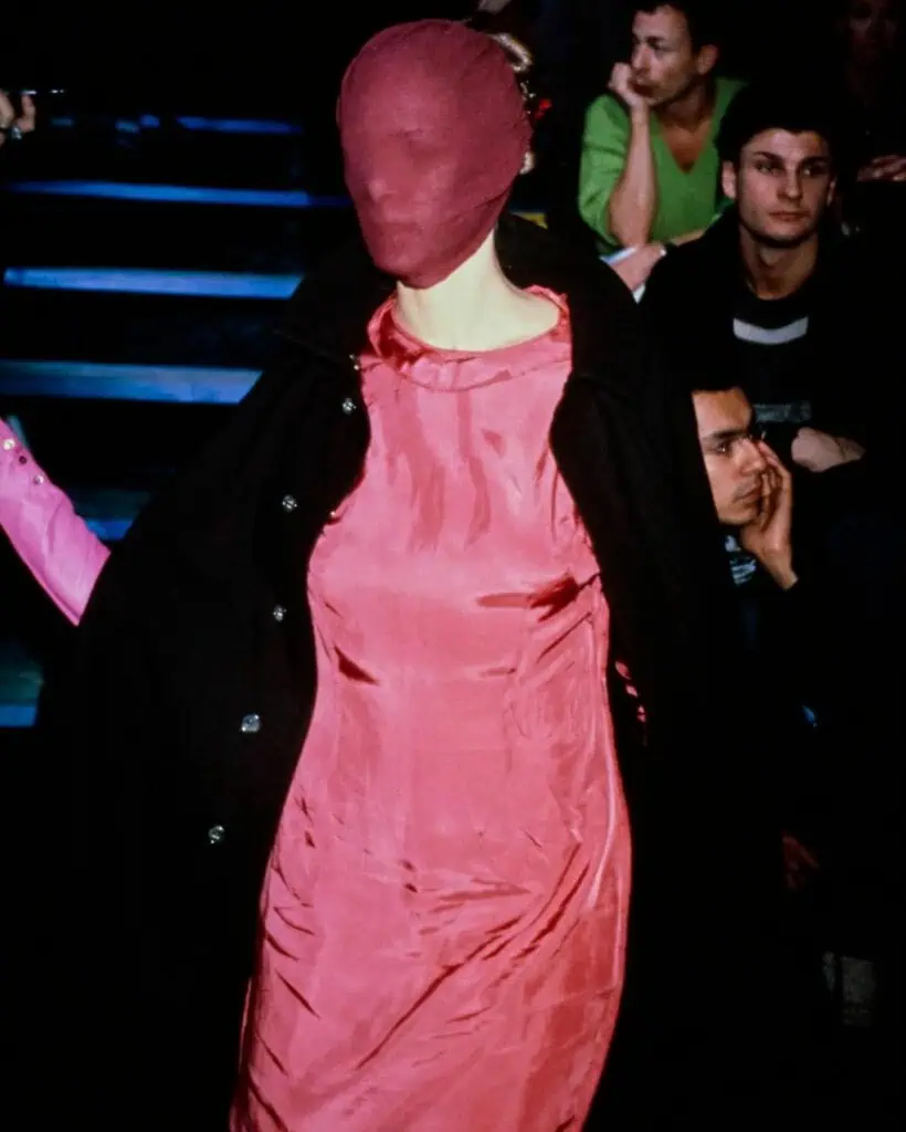A model walks on a runway wearing a shiny pink dress and a matching pink fabric mask covering their entire head, with a black coat draped over their shoulders. Audience members watch in the background.