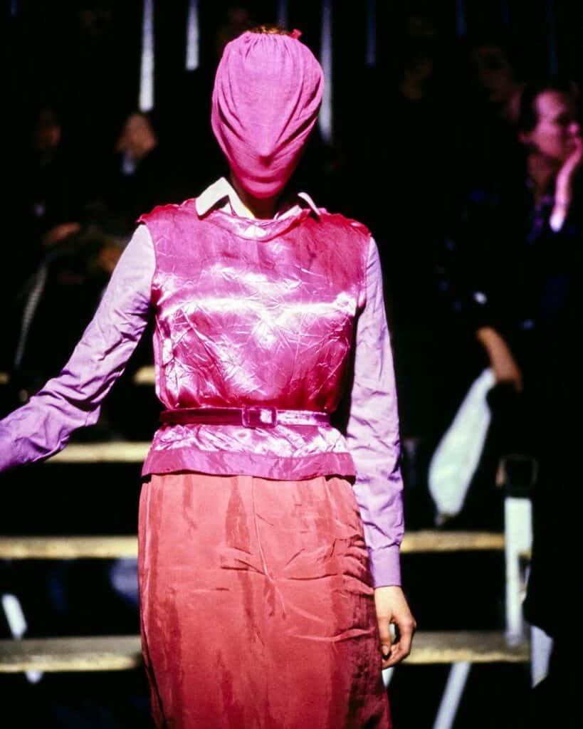 A model walks a runway wearing a shiny pink sleeveless top over a pink shirt and skirt, with a matching pink fabric covering their entire head. The background is dimly lit with people seated in the audience.