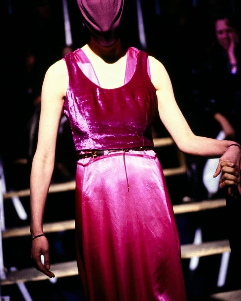A person wearing a shiny, sleeveless magenta dress and a matching fabric head covering walks on a runway, holding someone elses hand. The background is dimly lit with blurred audience members and steps visible.