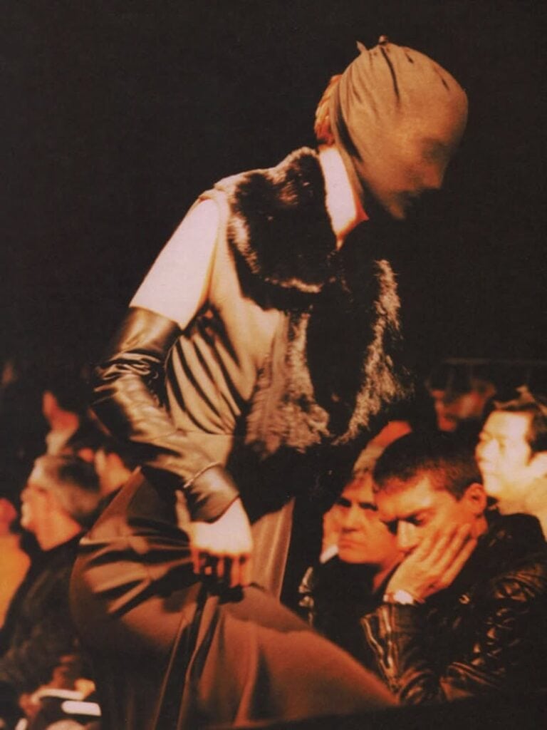 A fashion model wearing a sleeveless dress with dark gloves and a fur stole walks a runway with their head completely covered by a fabric mask; audience members watch closely in the background.