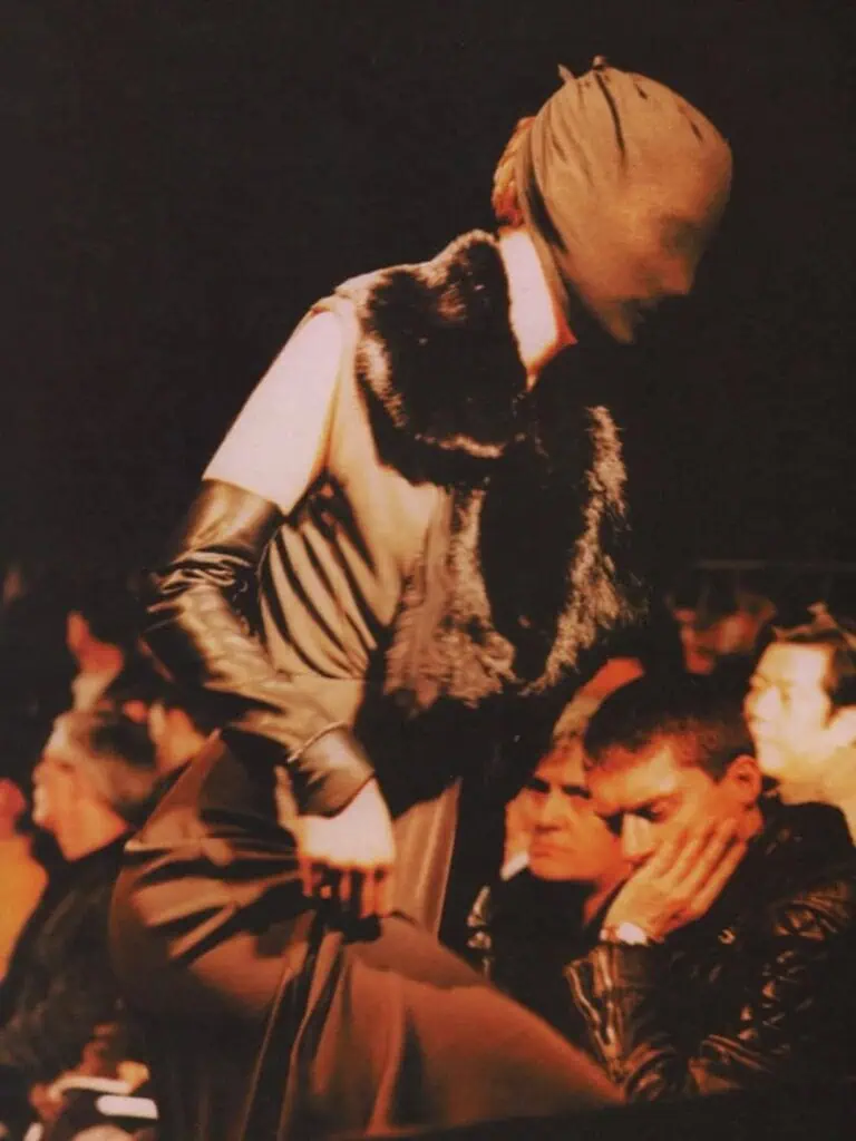 A fashion model wearing a sleeveless dress with dark gloves and a fur stole walks a runway with their head completely covered by a fabric mask; audience members watch closely in the background.