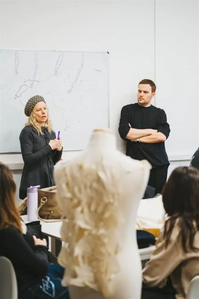 Two people stand in front of a classroom, one speaking and the other listening with arms crossed. A dress form with a ruffled fabric piece is in the foreground, and students are seated, watching them. A whiteboard is behind them.