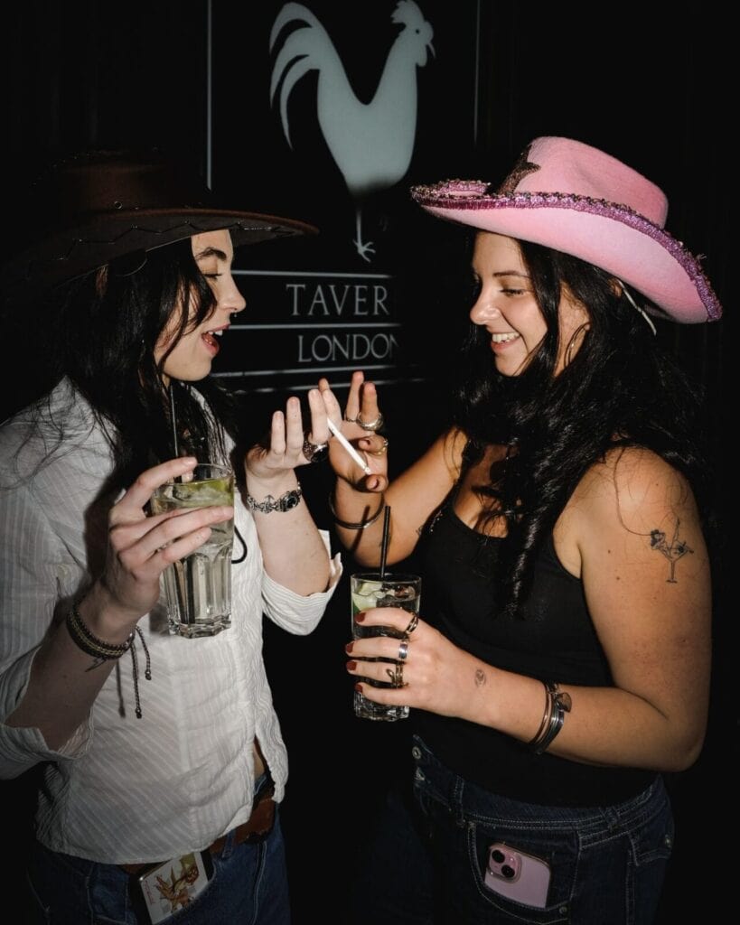 Two women in cowboy hats, one white and one pink, smile and talk while holding drinks and a cigarette in front of a Taver London sign with a rooster logo, sharing a fun Valentine moment in the dimly lit setting.