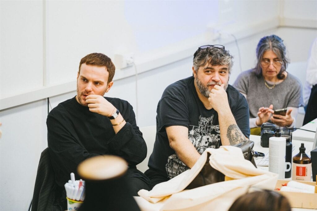 Three people sit at a table, two men in front with thoughtful expressions, one in a dark shirt and one in a graphic tee, and a woman behind them looking down while using a phone. Papers and items are on the table.