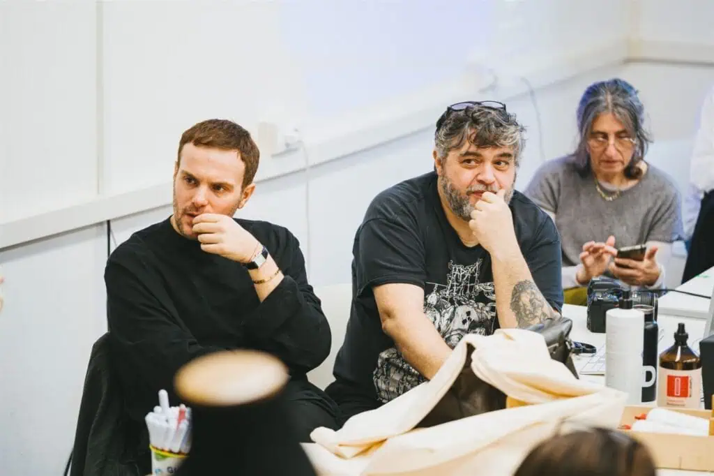 Three people sit at a table, two men in front with thoughtful expressions, one in a dark shirt and one in a graphic tee, and a woman behind them looking down while using a phone. Papers and items are on the table.
