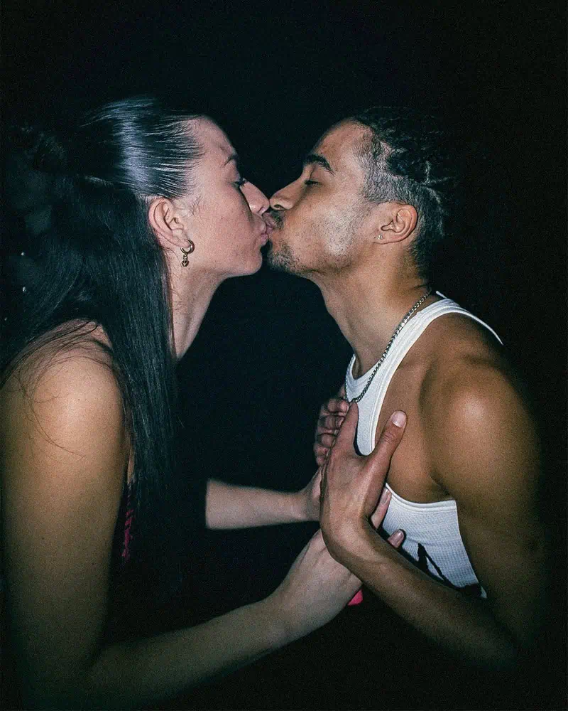 A woman and a man are standing close to each other, gently kissing on the lips. The scene is dimly lit, and the man has his hand on the womans shoulder while she holds his upper arm.