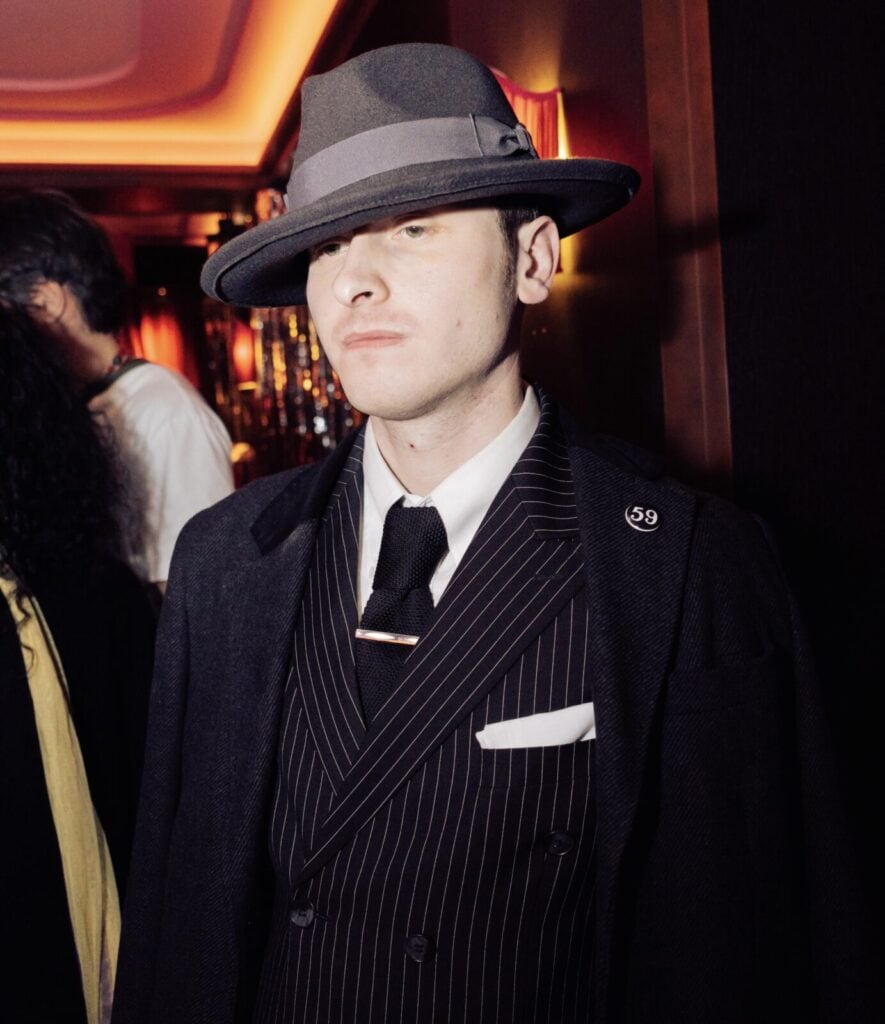 A man wearing a grey fedora hat, dark overcoat, and pinstripe suit with a tie stands indoors under warm lighting, evoking a vintage or 1920s gangster style. His face is partially shadowed by the hat.