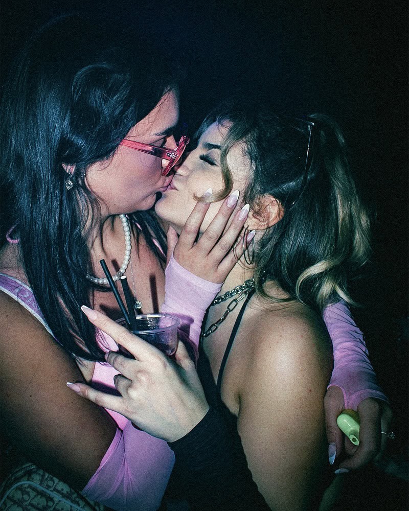 Two people are kissing in a dimly lit setting. One person holds a drink and wears pink glasses and gloves, while the other has blonde hair and wears a black strap top. Their embrace appears affectionate and intimate.