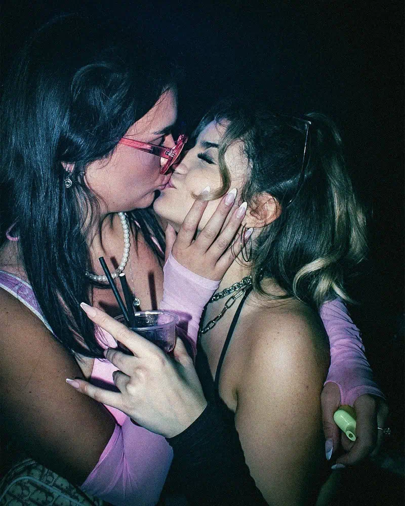 Two people are kissing in a dimly lit setting. One person holds a drink and wears pink glasses and gloves, while the other has blonde hair and wears a black strap top. Their embrace appears affectionate and intimate.