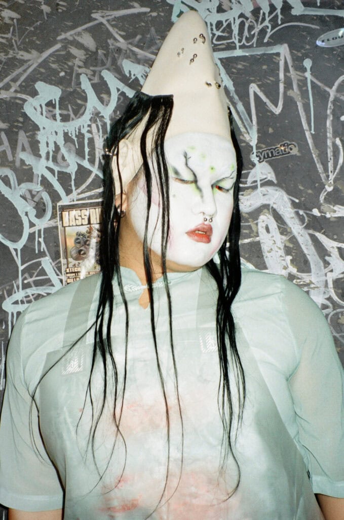 A person with elaborate white face makeup, black drawn lines, green accents, fake piercings, and long black hair wears a tall, pointed headpiece and a sheer mint top, standing against a graffiti-covered wall.