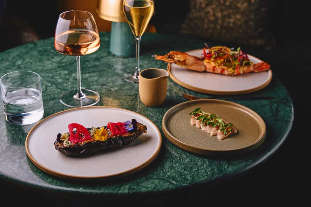 A green marble table set with three artfully plated dishes, glasses of wine and champagne, a cup, and a glass of water; the dishes include garnished eggplant, seafood, and sliced meat with microgreens.