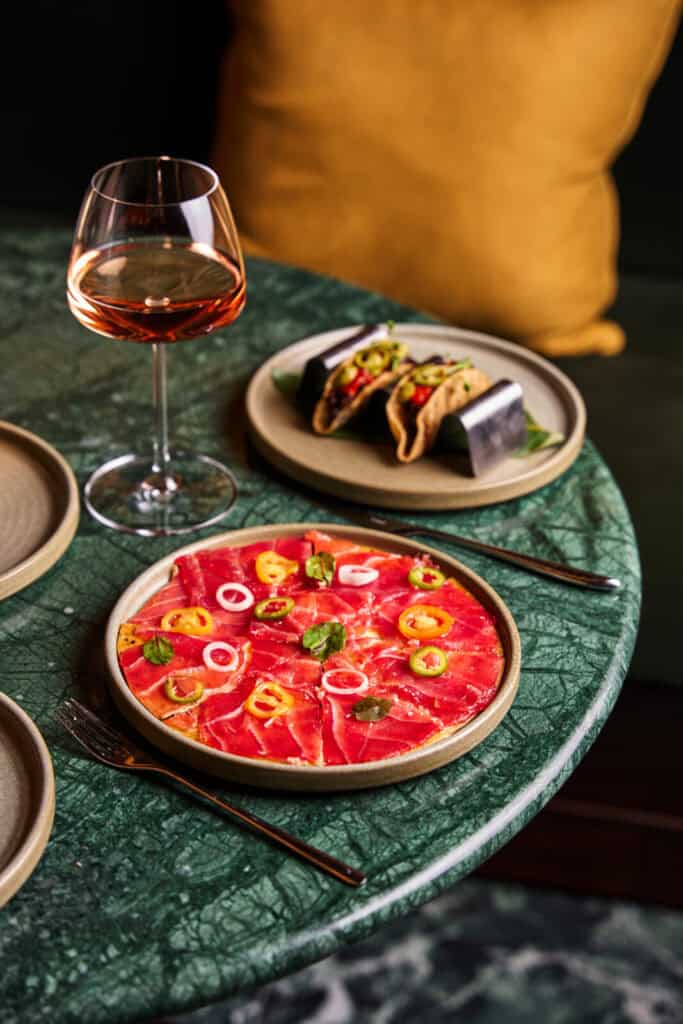 A round green table set with a plate of thinly sliced tuna carpaccio garnished with herbs and peppers, a plate holding two tacos in a metal stand, and a glass of rosé wine. A mustard yellow pillow is in the background.