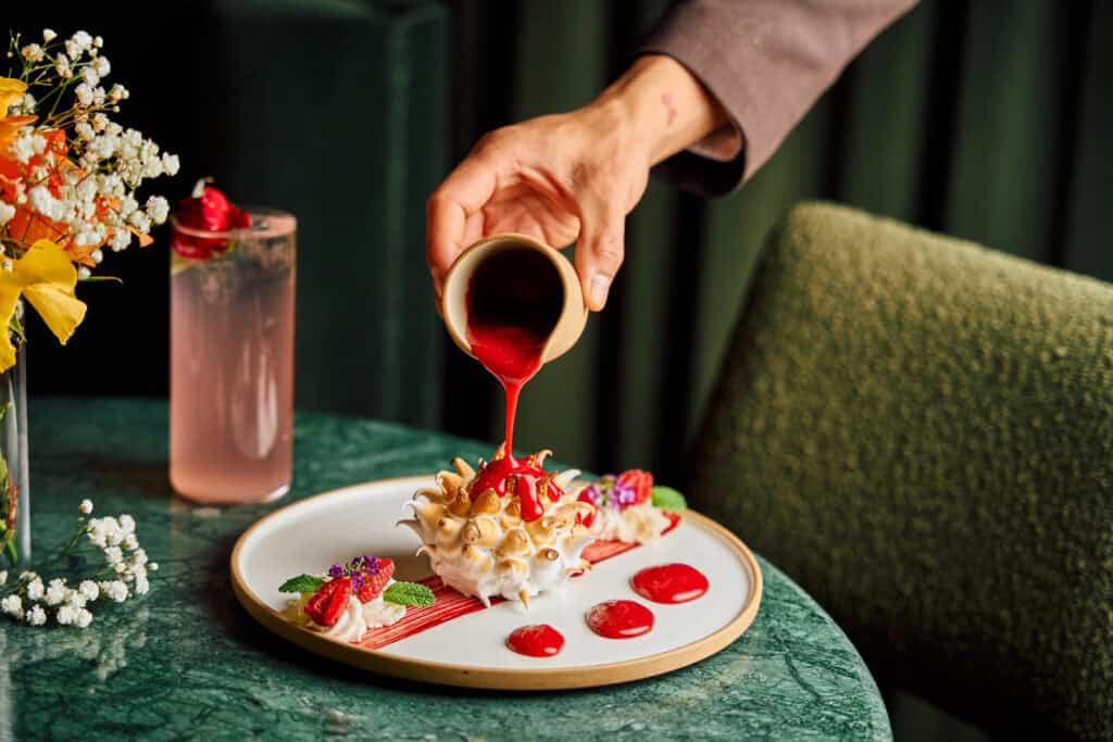 A hand pours vibrant red sauce onto a plated dessert topped with toasted meringue, strawberries, and mint, with a pink drink and flowers on a green marble table.