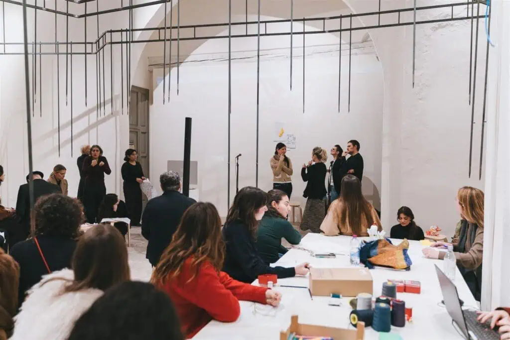 People are gathered in a bright room around tables, some sitting and working, others standing and speaking at the front. The space has white walls and minimal decor, with materials and papers spread out on the tables.